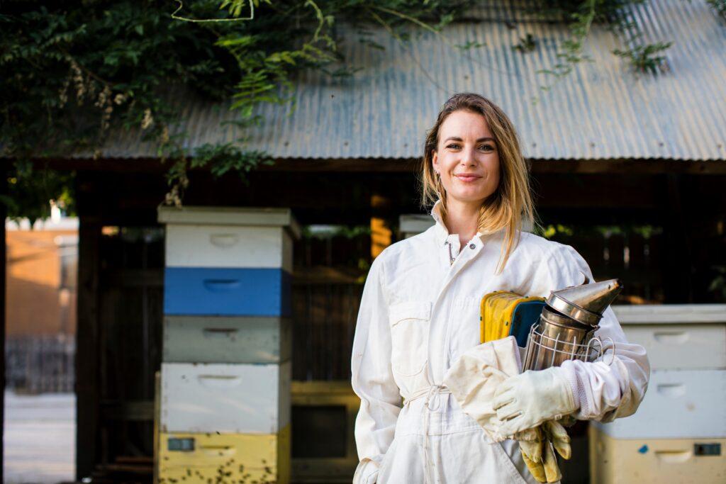 Eine lächelnde Imkerin in Schutzkleidung steht vor ihren Bienenstöcken, symbolisiert die nachhaltige Gewinnung von Bienengift für die Herstellung von Heilsalben.