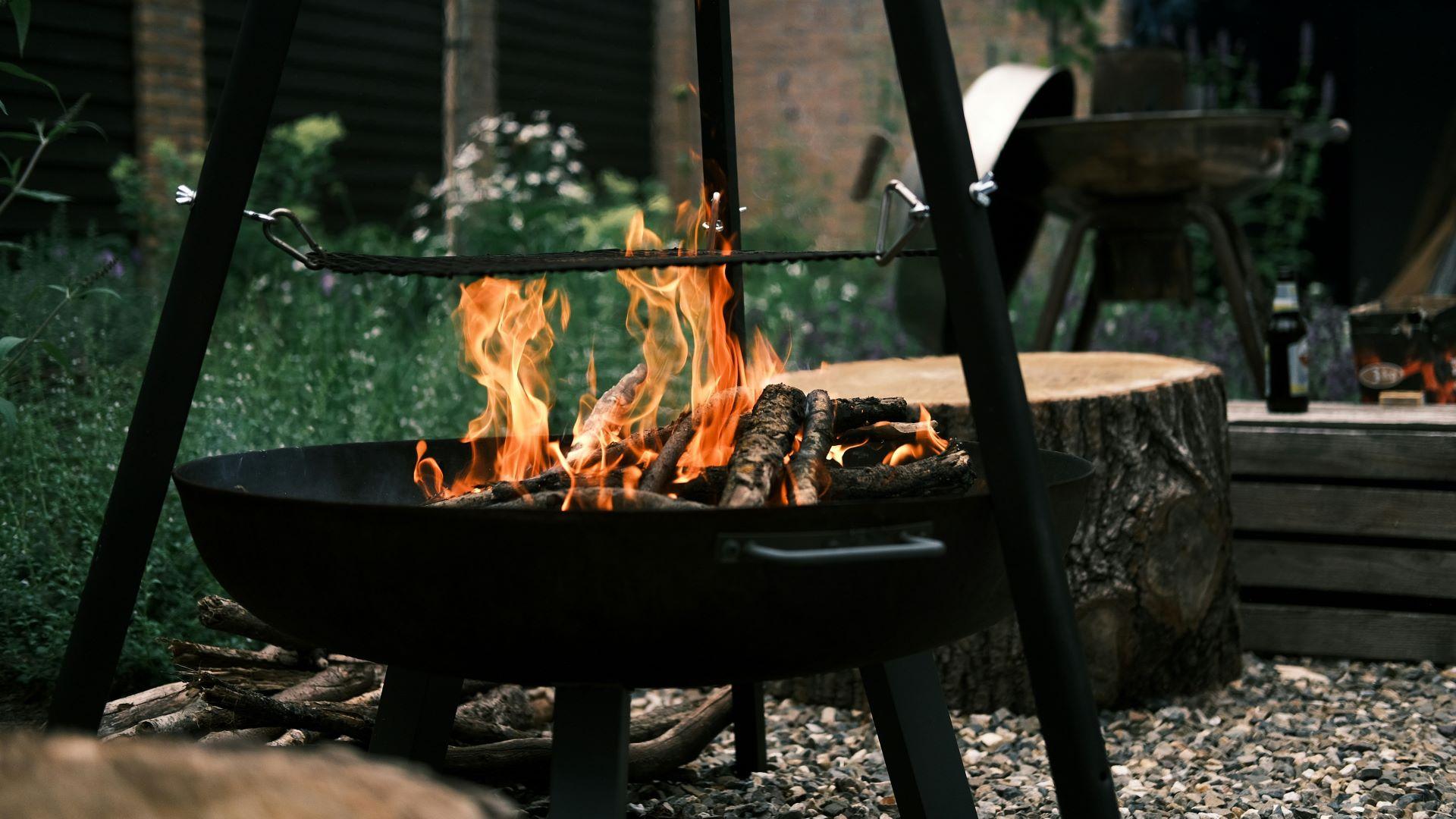 Moderne Feuerschale mit brennenden Holzscheiten im Garten, umgeben von rustikaler Einrichtung und Pflanzen.